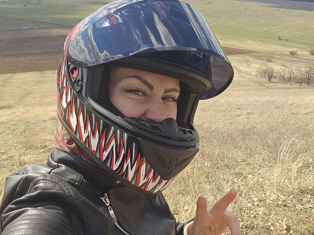 JuliaaCherry mit einem glänzenden, rot-weißen Motorradhelm, einer schwarzen Lederjacke und einem zwinkernden Gesicht, die eine "rock on"-Handgeste macht und in einem grasbewachsenen Feld steht..