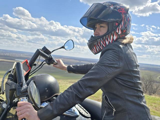 JuliaaCherry, vestindo uma jaqueta de couro preta e um capacete com padrão vermelho e branco, pilota uma motocicleta com um capacete preto na mão direita, sob um céu azul e nublado..
