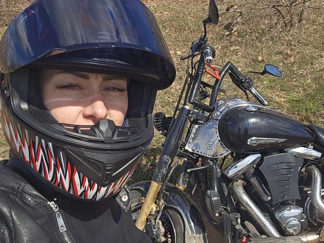 JuliaaCherry, con un casco nero a motivi di denti di squalo, sorridente, indossa una giacca di pelle nera e sta in piedi accanto a una motocicletta nera lucida con dettagli cromati..