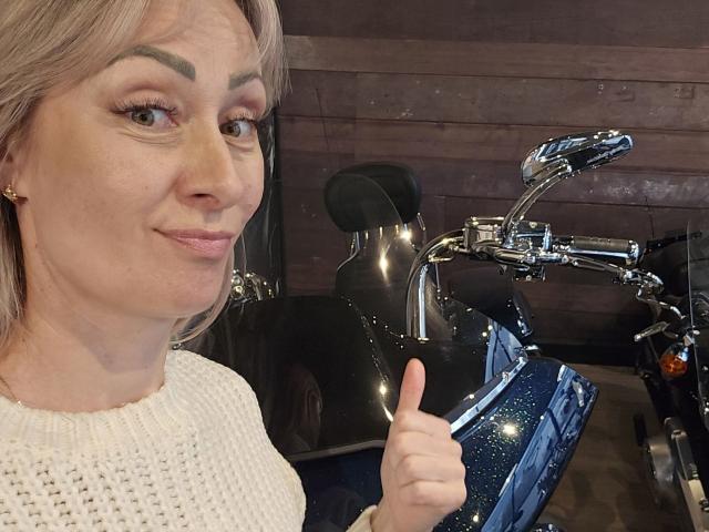 JuliaaCherry, com cabelo loiro, vestindo um suéter branco de tricô, dando um joinha ao lado de uma motocicleta preta brilhante com detalhes cromados..
