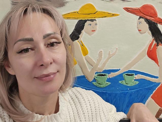 JuliaaCherry mit blonden Haaren und einem weißen Strickpullover, lächelnd vor einem Gemälde von zwei Frauen mit Sonnenhüten, roten Kleidern und grünen Tassen auf einem blauen Tisch..
