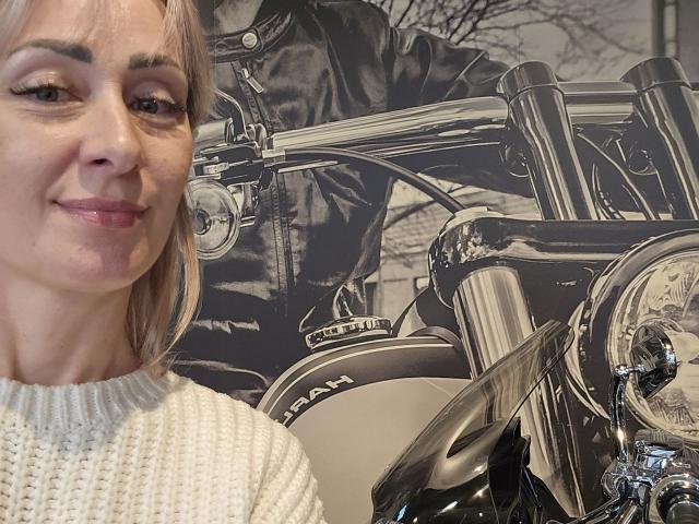 JuliaaCherry, uma mulher loira, sorrindo, vestindo um suéter branco de tricô, com uma motocicleta cromada brilhante em um fundo preto e branco, foto. Os seios não estão visíveis, mas ela tem um rosto bonito e uma vibe relaxada..