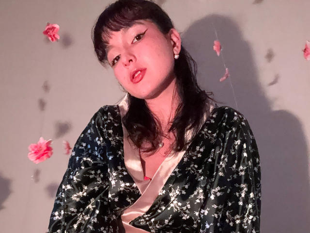HairyValeri with dark hair, wearing a floral green kimono, red lips, and eyeliner, looking slightly up, shadow on wall, pink flower decorations, soft light, slightly messy.