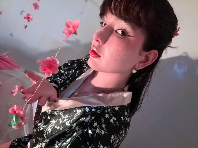 HairyValeri with dark hair, wearing a black floral top, white collar, and pearl earrings, looking up with pink flower petals floating around, red lipstick, and a serious expression.