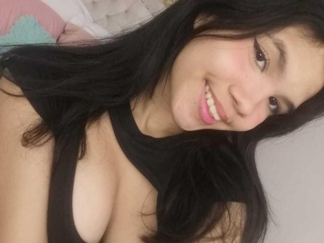MidnightHarpy with long black hair, smiling at the camera, showing her tits in a black top, pink lipstick, and light brown skin, lying on a bed.