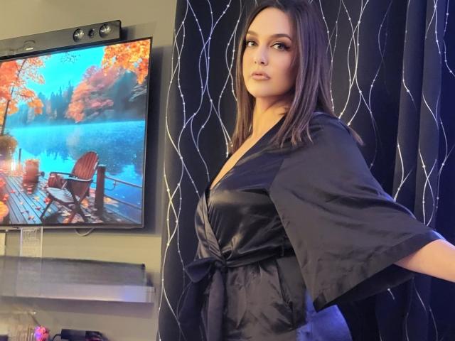 SelinaKylla with medium brown hair, wearing a black satin robe, standing in front of a TV showing a colorful autumn scene, looking to the side with a neutral expression.