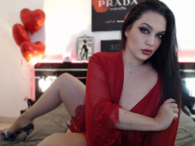 SelinaKylla with red lace robe, revealing tits, sitting on bed, legs crossed, wearing black heels, long black hair, red lipstick, and red balloons in background.