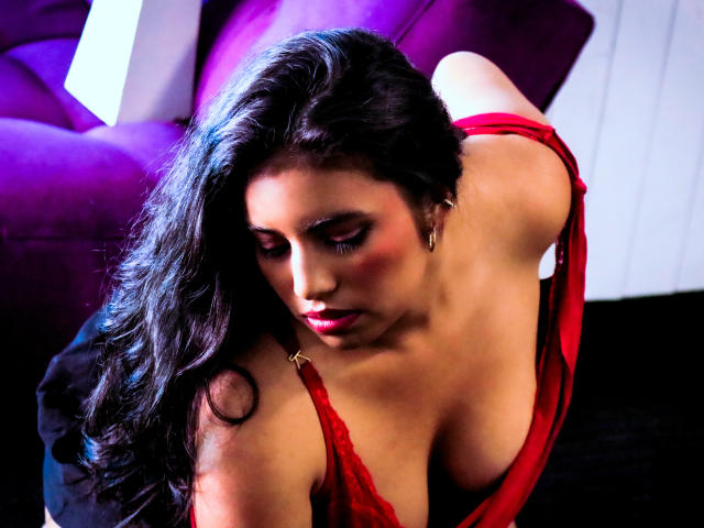 DaphneVilla with long black hair, red lingerie, and visible large tits, leaning forward, eyes closed, pink lipstick, and a purple couch in the background.