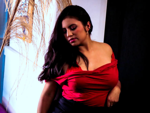 DaphneVilla with long dark hair, red off-shoulder top, showing tits, black skirt, looking down, black curtain, puffy pink lips, subtle makeup, confident, sexy, warm light.