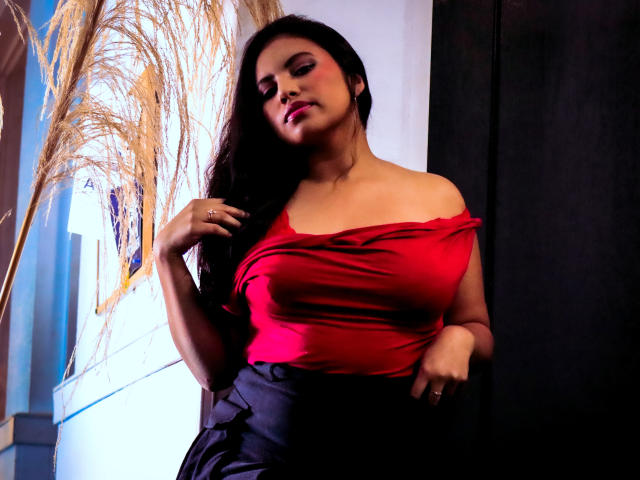 DaphneVilla with long black hair, red off-shoulder top, black skirt, and ring, touching hair, looking down, soft light, black curtain, pampas grass in background.