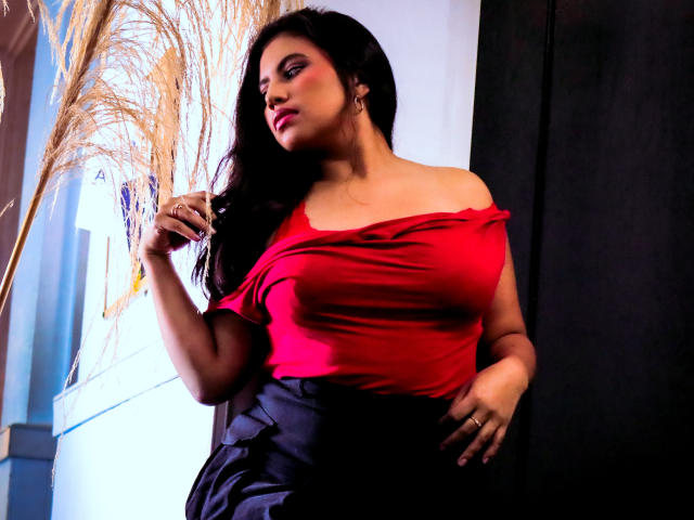 DaphneVilla with long black hair, red off-shoulder top, showing her tits, black skirt, holding pampas grass, standing against dark wall, looking to the side with pink lipstick.