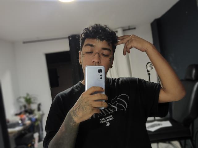 SteveArt with curly black hair, glasses, and tattoos, holding a white iPhone, taking a mirror selfie, wearing a black shirt, hand touching head.