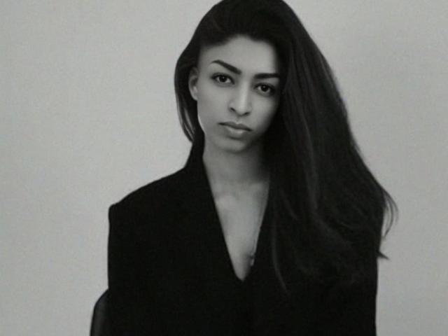 BlackMoonTS with long dark hair, serious expression, wearing a black jacket, showing a slight cleavage, black-and-white photo, looking directly at the camera.