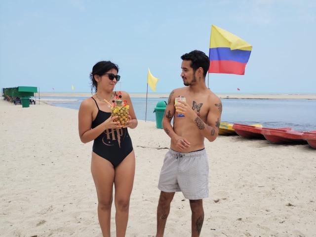 LailaAsher with black swimsuit holding fruit, medium tits, and dark hair, stands next to shirtless LailaAsher with gray shorts, tattoos, and black hair, holding drink on beach.