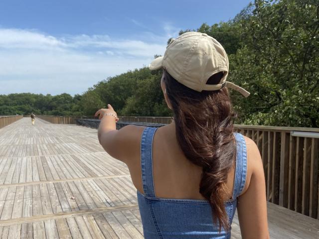 Laila Asher met een vlecht, een beige pet en een blauwe spijkerjurk, wijzend vooruit op een houten brug, met haar rug naar de camera, zonnige dag, bos op de achtergrond..