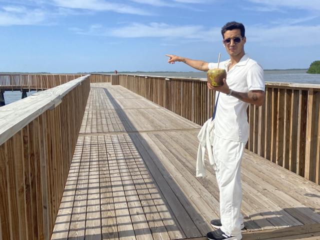 LailaAsher stands on a wooden boardwalk, pointing ahead with a green drink in hand. She's wearing a white polo and pants, with sunglasses and a white bag.