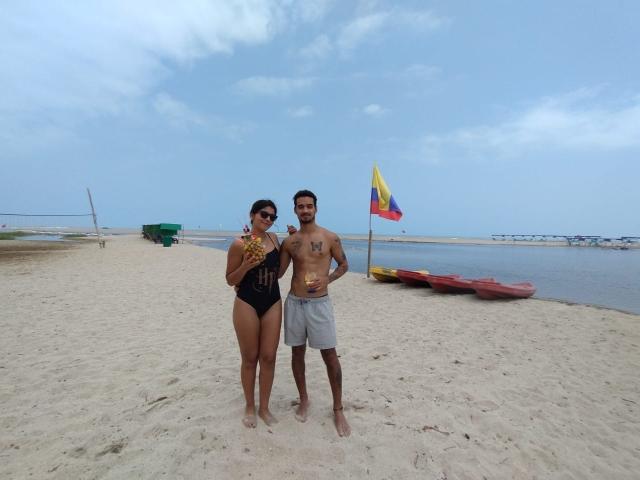 LailaAsher, com seios médios, usando um maiô preto de peça única, segurando uma bebida amarela, está ao lado de LailaAsher sem camisa, com o peito tatuado e shorts cinzas..