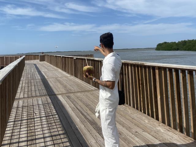 LailaAsher in white clothes, holding a coconut, pointing at the water, standing on a wooden boardwalk with railings, under a blue sky with wispy clouds.
