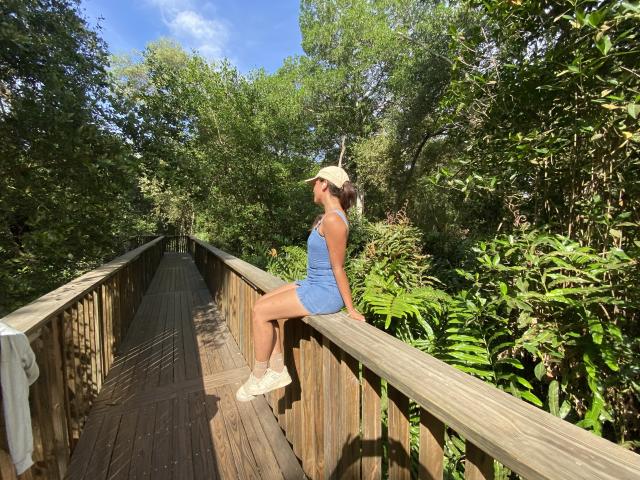 LailaAsher, wearing a blue dress and white cap, sits on a wooden bridge with her ass on the railing, surrounded by green foliage and blue sky.