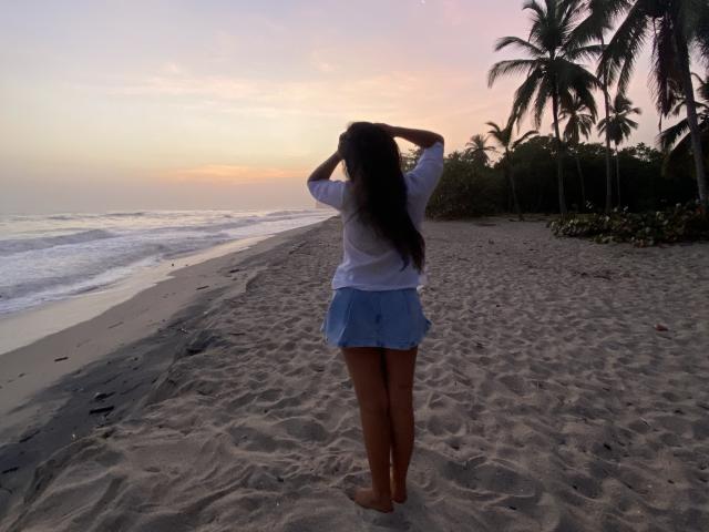 Laila Asher está de pie en una playa arenosa, de espaldas a la cámara, con las manos en su largo cabello oscuro, vistiendo una camisa blanca y una falda azul, atardecer, palmeras, olas del océano..