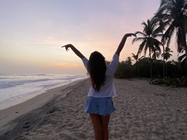 LailaAsher stands on a beach, arms raised, wearing a white shirt and blue skirt, with long brown hair, facing the sunset, palms on the right, ocean on the left.