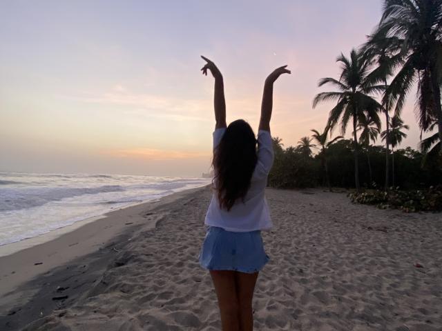 Laila Asher, con los brazos levantados, vestida con una camisa blanca y una falda azul, de pie en una playa arenosa al atardecer, de espaldas a la cámara, con palmeras a la derecha..