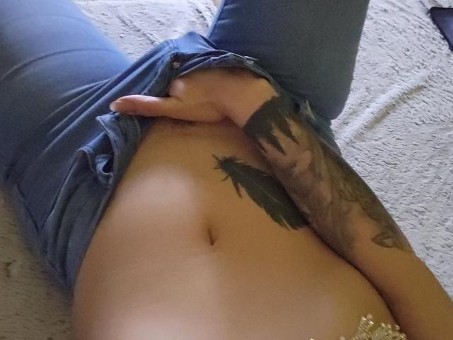 TheSunshine's hand is pulling down blue jeans to reveal a tattooed arm and their bare, flat, tan-lined belly with a visible navel. TheSunshine's thumb is slightly curled.