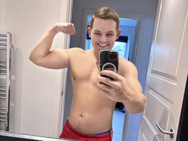 rasmusboy, shirtless, flexing bicep, smiling, holding phone, red shorts, mirror selfie, white door, gray wall, towel rack on left, brown hair, fair skin, muscular, short hair.