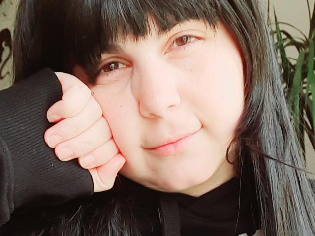 IvyBloom with black hair, bangs, and a nose ring, smiling slightly, wearing a black shirt, resting her chin on her hand, showing pale skin and brown eyes.