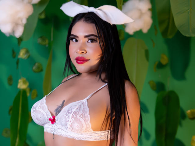 OliviGrace with black hair, white lace bra, red bow, white headband, and red lipstick, showing her tits, tattoo below, green background with white flowers.