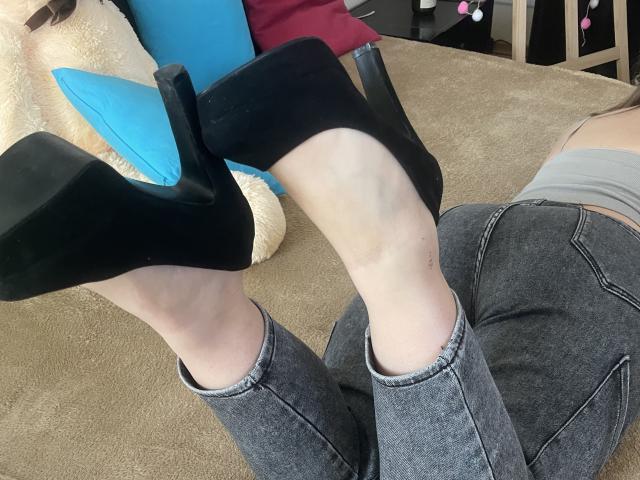 Las piernas de Madrinia están elevadas, lleva tacones negros altos y pantalones vaqueros grises. Su piel pálida es visible. Está acostada sobre una alfombra beige, con un cojín azul en el fondo..