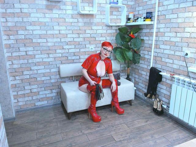 Lackengel-hot, with red latex outfit, big tits, red boots, holding black flogger, sitting on white bench, wearing glasses, in brick-walled room, wooden floor, potted plant, radiator.