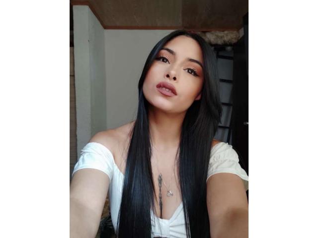 EmilyVv with long black hair, pouty lips, and a white off-shoulder top, showing her tits slightly in a selfie. She's wearing a necklace with a small pendant.
