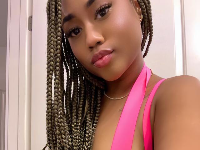 BriannaBrave with braided hair, pink halter top, glossy brown skin, and pink lips, wearing a gold necklace, looking at the camera with a slight smile.