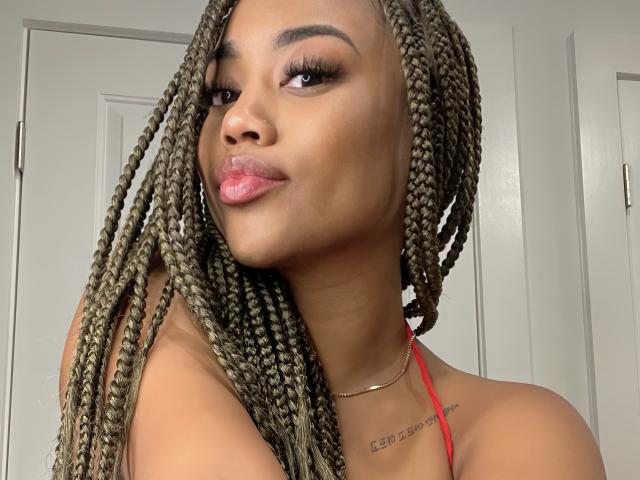 BriannaBrave, avec sa peau brune, ses longues tresses blondes, ses lèvres roses pulpeuses, un maquillage discret, et un haut rouge en chaîne, sourit légèrement à l'appareil photo. Elle porte un collier fin en or..