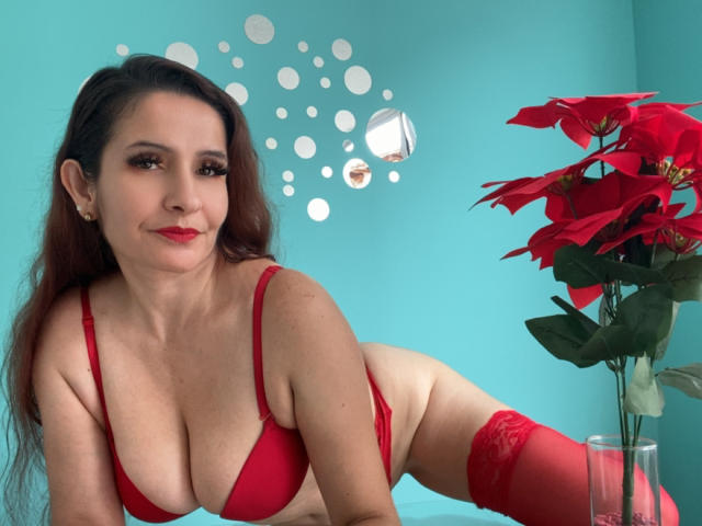 Charis with red bra and panties, red garter, and red poinsettia, has large tits, long brown hair, and red lipstick, leaning forward with a slight smile.