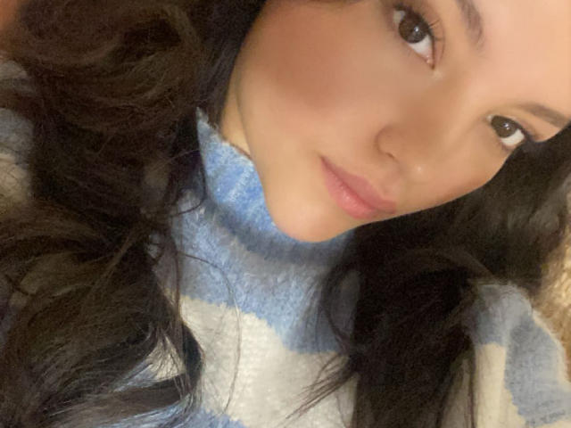 IssaPuasini has long, wavy black hair, brown eyes, and a cute, slightly parted pink lip, wearing a blue and white striped sweater. Her ass is not visible.