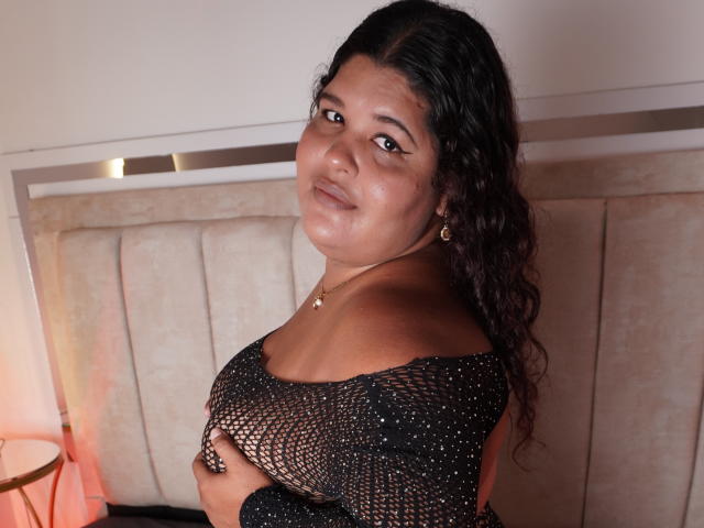CharlotSuny, met krullend donker haar, een medium bruine huid en kleine gouden oorbellen, draagt een zwarte kanten top, houdt haar borsten vast en kijkt met een lichte glimlach naar de camera..