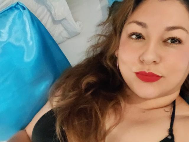 SakotoDika, com batom vermelho, cabelo castanho ondulado, sutiã preto e brincos de argola, sorrindo para a câmera. Seus seios estão visíveis, mostrando um tamanho de busto moderado..