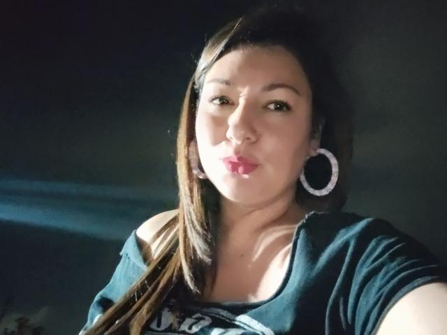 SakotoDika mit langen, braunen Haaren, die ein grünes Hemd trägt, mit Ohrringen und pinkem Lippenstift, zieht die Lippen zusammen und schaut in die Kamera. Ihre Brüste sind leicht sichtbar..