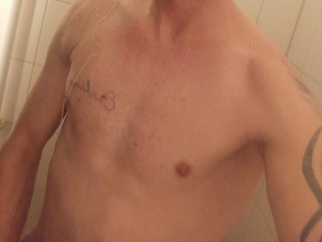 MilfAnaLyzer-hot, con il petto nudo e piccoli capezzoli, un tatuaggio sul lato sinistro e un tatuaggio scuro sul braccio destro, sotto la doccia, con l'acqua che bagna la pelle..