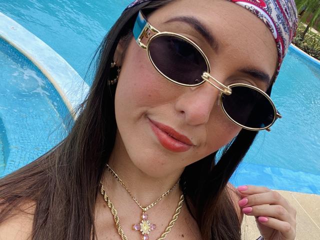 YejSusen, con i capelli castano scuro, indossa una sciarpa rossa con motivi, occhiali da sole aviator dorati, rossetto rosa e una collana con un fiore rosa, sorridendo accanto alla piscina blu..