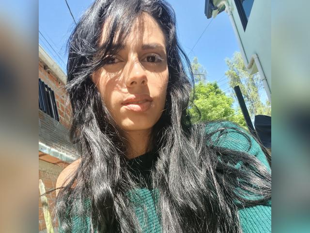 AnaisHilton has long, wavy black hair, brown eyes, and a serious expression, wearing a green rib-knit top, with sunlight highlighting her tan skin and subtle makeup.
