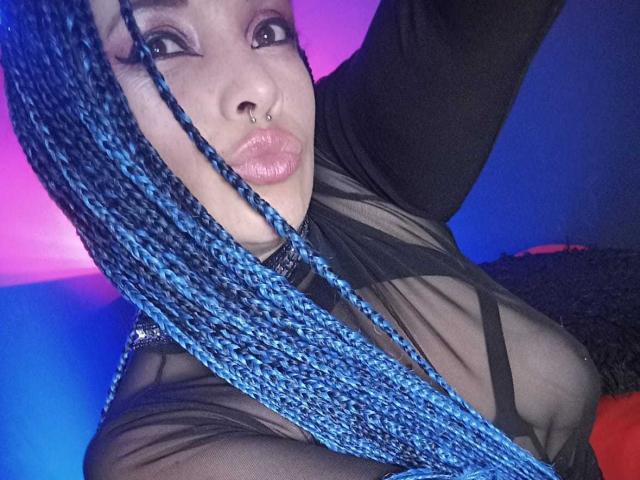 DalilaMusa with blue braided hair, black sheer top, nose ring, and pouty lips, showing her tits and looking at the camera with a confident expression.