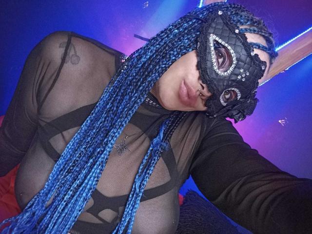 DalilaMusa with blue braids, black mask, and sheer top, showing her tits through the fabric. She's got a nose ring and a necklace. Blue and purple lights in background.