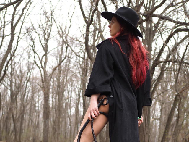 MelanyTessk with long red hair, black hat, and coat, holding a black leather strap, standing in a leafless forest, showing her bare ass with a black thong.