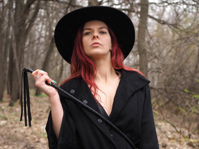 MelanyTessk with red hair, black hat, and coat, holding a black whip, standing in a forest, showing her tits, looking confident. Her green nails are visible.