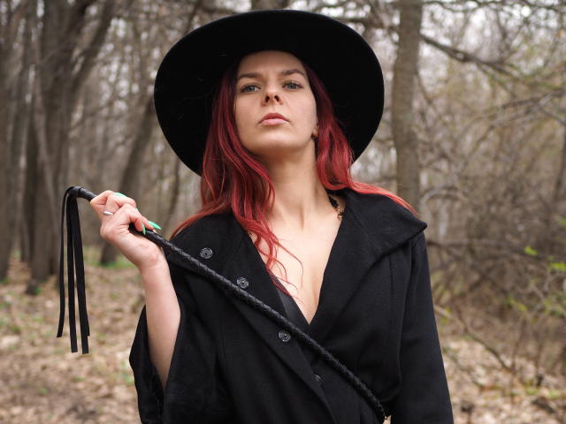 MelanyTessk with red hair, black hat, and black coat, holding a black leather flogger, looking confident in a forest. Boobs slightly visible under coat.