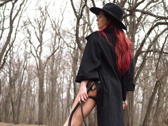 MelanyTessk, con cabello rojo, sombrero negro y abrigo negro, sosteniendo correas negras, de pie en un bosque, mostrando su trasero desnudo a través del abrigo..