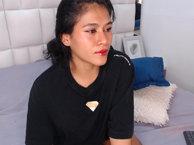 AnnieDarling, a cute Asian woman with dark hair in a ponytail, wearing a black shirt with a heart, red lipstick, and subtle makeup, sits on a bed.
